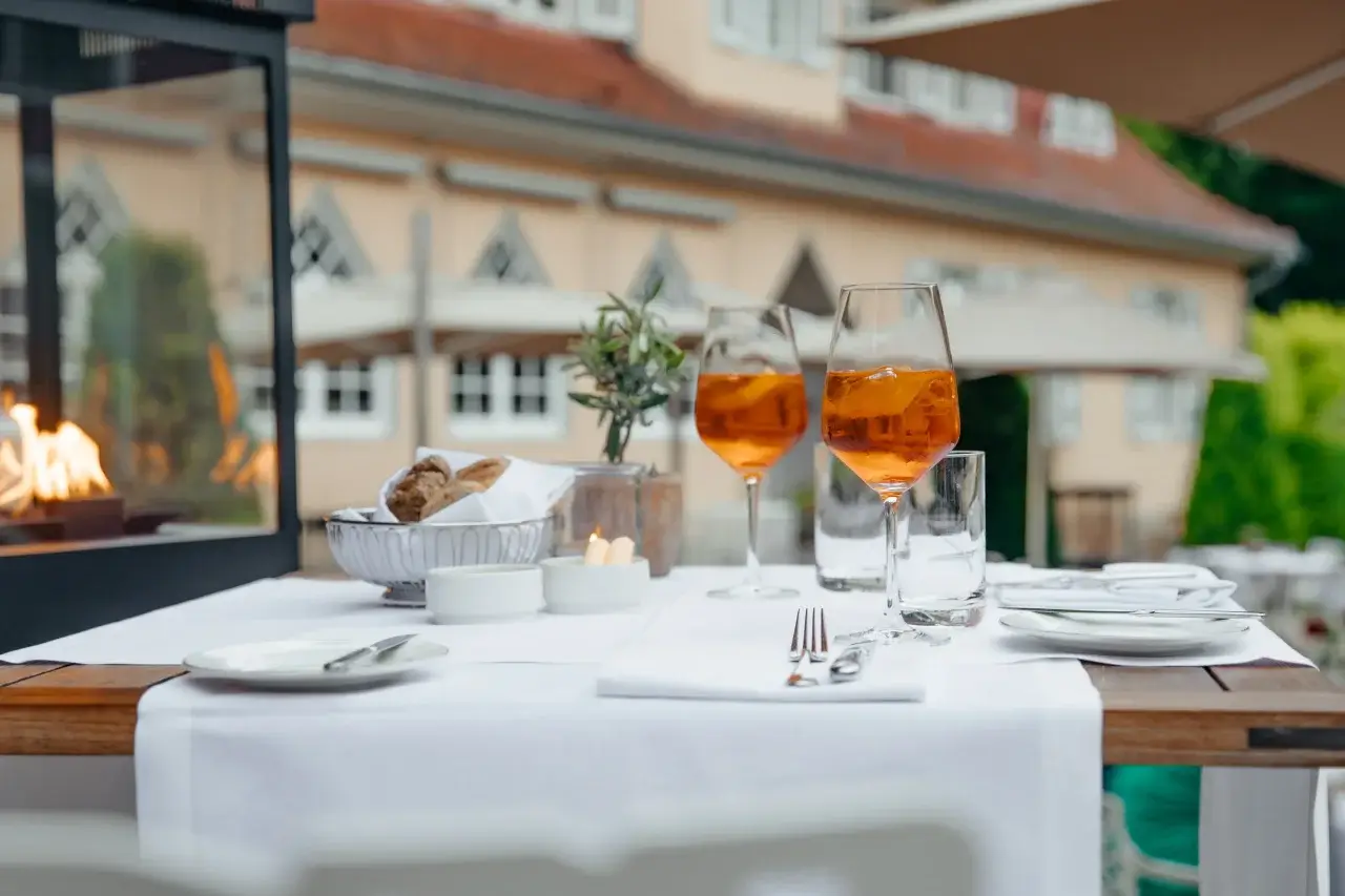 Gedeckter Tisch auf einer Terrasse eines Restaurants mit Aperol Spritz und Brotkorb, im Hintergrund das charmante Waldhotel und ein Kamin.