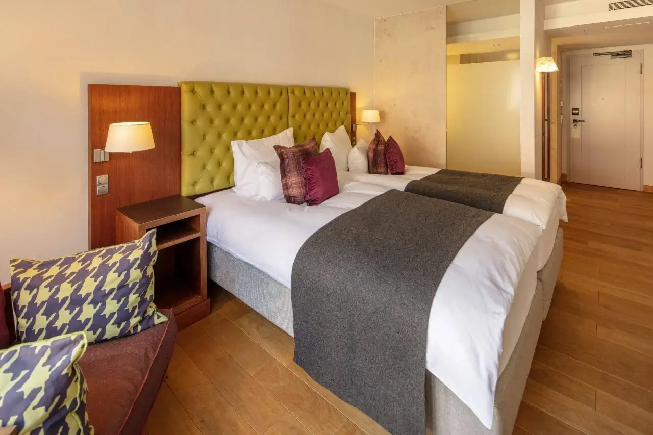 Modernes Hotelzimmer mit zwei Einzelbetten, gelbem Kopfteil, lila Kissen und gemütlicher Sitzecke.