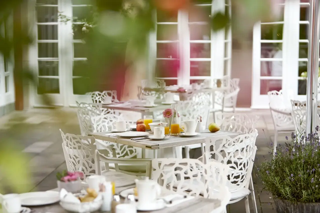 Eine stilvolle Gartenterrasse mit weißen Stühlen und gedeckten Tischen für ein Frühstück im Freien.