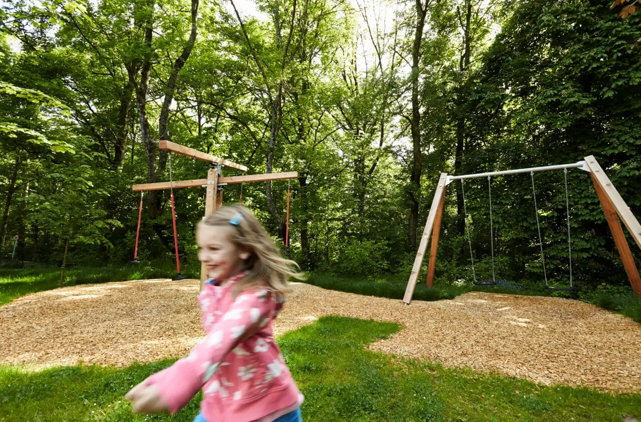 Ein kleines Mädchen rennt lachend über den Spielpaltz des Waldhotels mit verschiedenen Schaukeln.