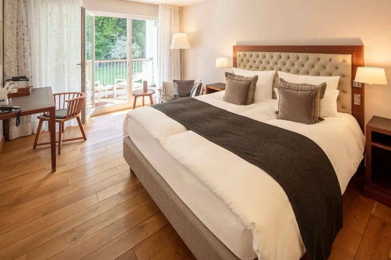 Gemütliches Hotelzimmer mit großem Doppelbett, Holzboden, Schreibtisch und Zugang zu einem Balkon mit weißen Stühlen mit Blick ins Grüne.