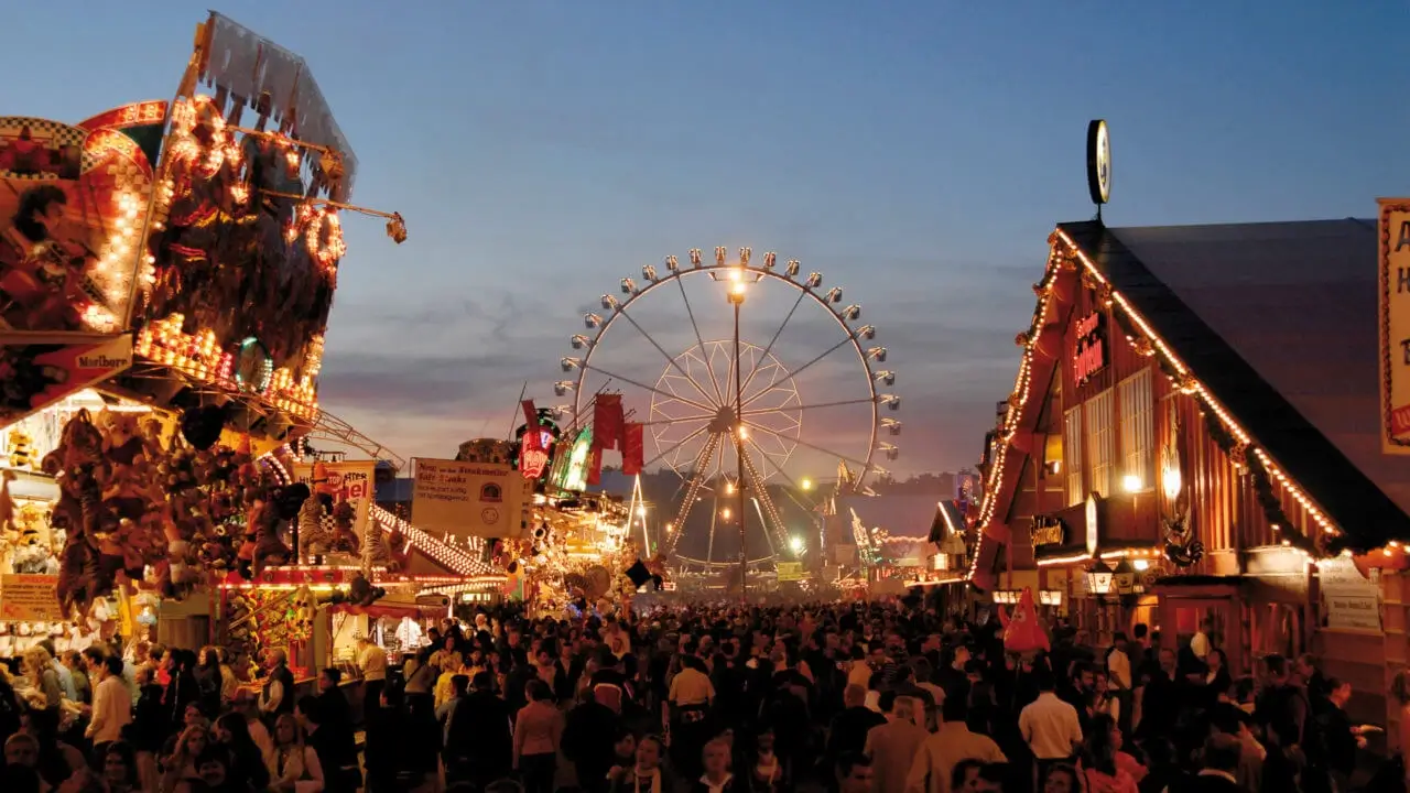 Der Cannstatter Wasen bei Abenddämmerung mit Hütten, Fahrgeschäften und Riesenrad.