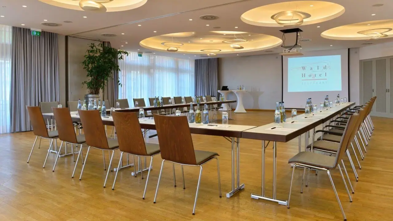 Moderner Tagungsraum im Waldhotel Stuttgart mit U-förmiger Tischanordnung, Holzboden, hellen Vorhängen und einer großen Leinwand.