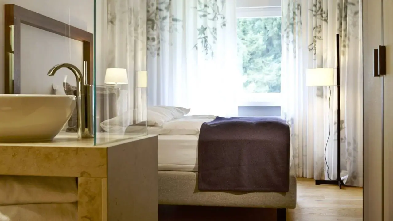 Modernes Hotelzimmer mit einem gemütlichen Bett, einer stilvollen Waschtischarmatur und großen Fenstern, die von zarten, bedruckten Vorhängen umrahmt sind.