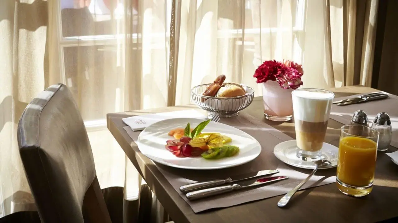 Ein gedeckter Tisch in einem Hotel mit Frühstück in Stuttgart mit einem Teller frischem Obst, einem Latte Macchiato, einem Glas Orangensaft, einer Serviette mit Hotellogo, einem Blumenstrauß und einer Schale mit Brötchen und Brezeln.