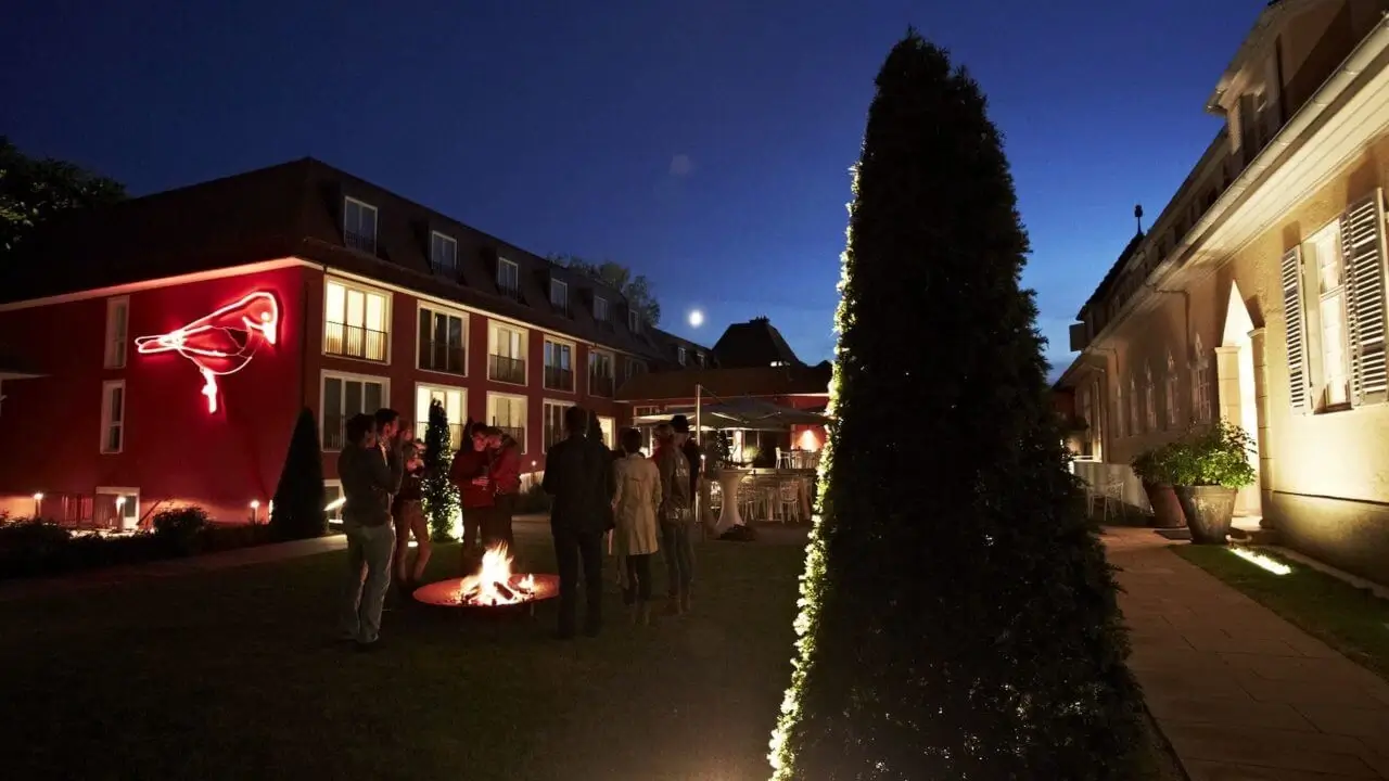 Das nächtliche Hotelgelände mit beleuchteten Gebäuden, einer Gruppe von Menschen einer Firmenfeier in Stuttgart, die um ein Lagerfeuer versammelt sind, und einem roten Vogelbild an der Hauswand.