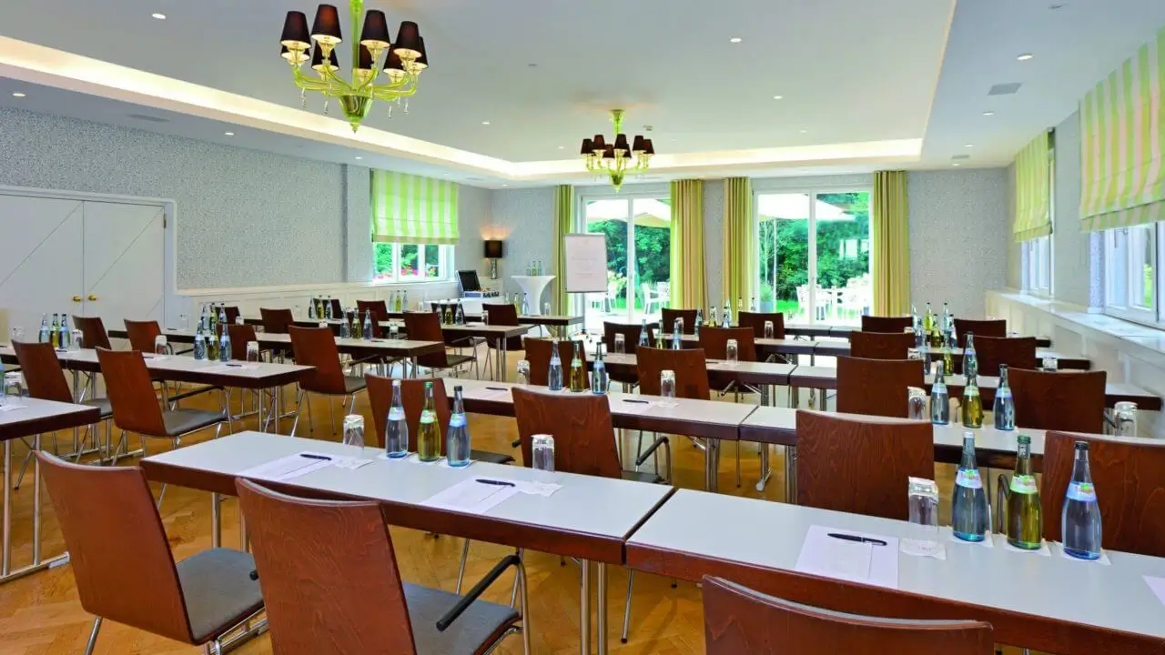 Ein heller Konferenzraum im Waldhotel Stuttgart mit Tischen und Stühlen, ausgestattet mit Wasserflaschen, Schreibutensilien und einem Flipchart, sowie großen Fenstern, die viel Tageslicht hereinlassen.