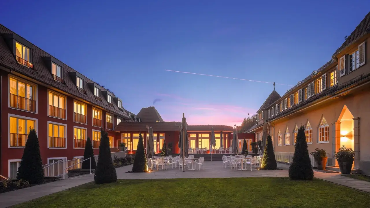 Stimmungsvolle Außenansicht eines stilvollen Hotels bei Sonnenuntergang, mit beleuchteten Fenstern und gemütlicher Terrasse.