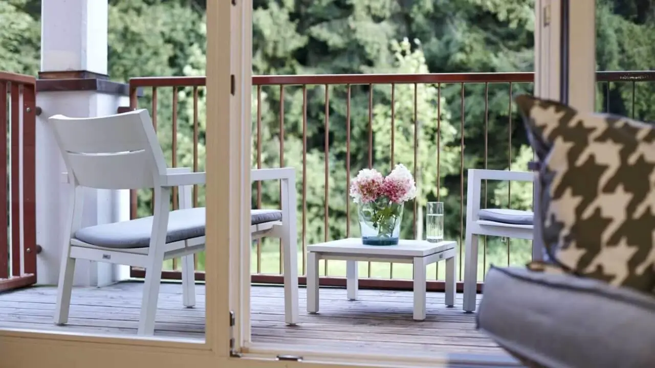 Ein gemütlicher Balkon im Waldhotel Stuttgart mit weißen Gartenmöbeln, einem kleinen Tisch mit Blumen und einem Glas Wasser, mit Blick auf die umliegende Natur.